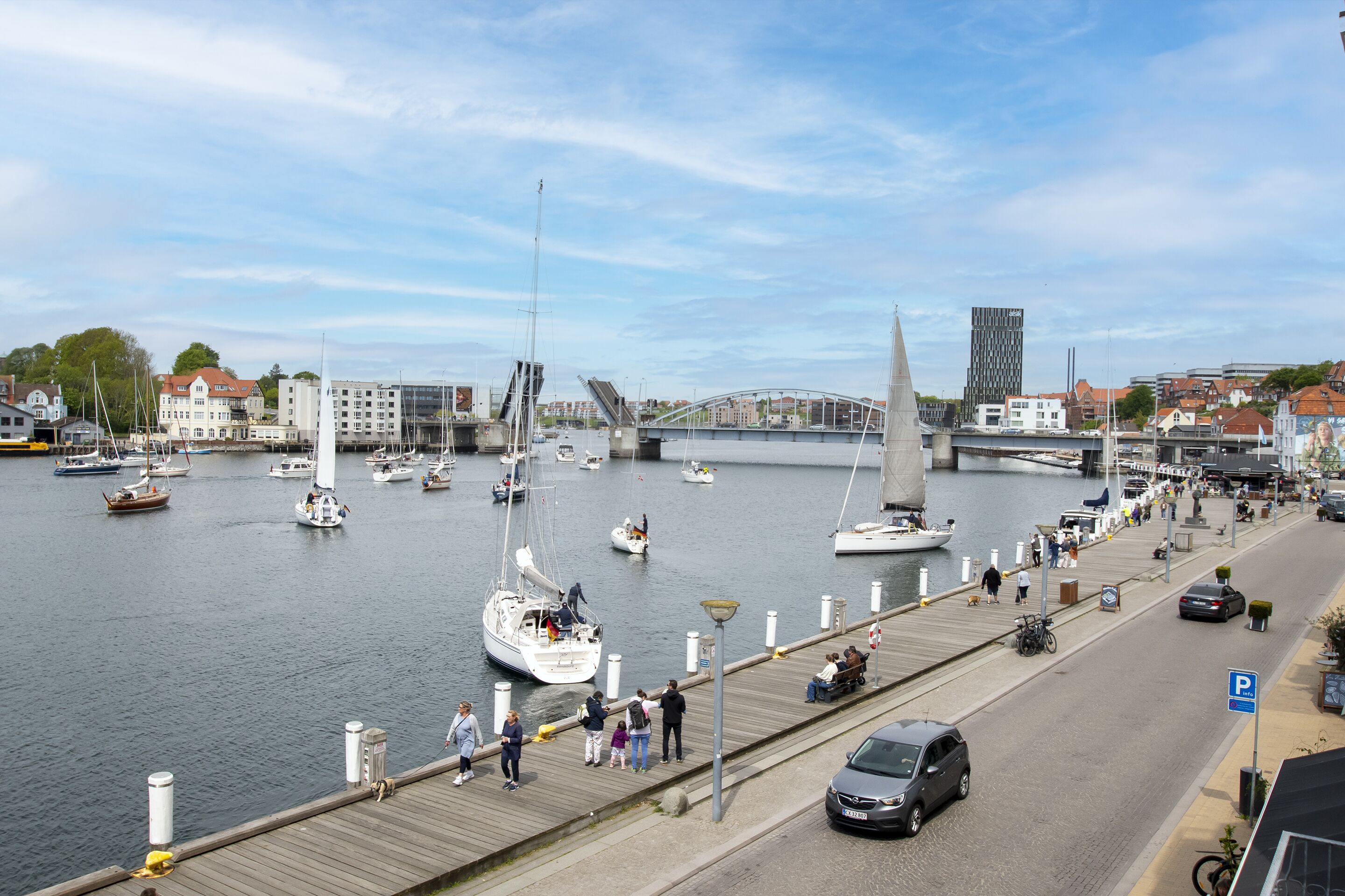 A marina with boats, a pedestrian walkway, and a bridge in the background.