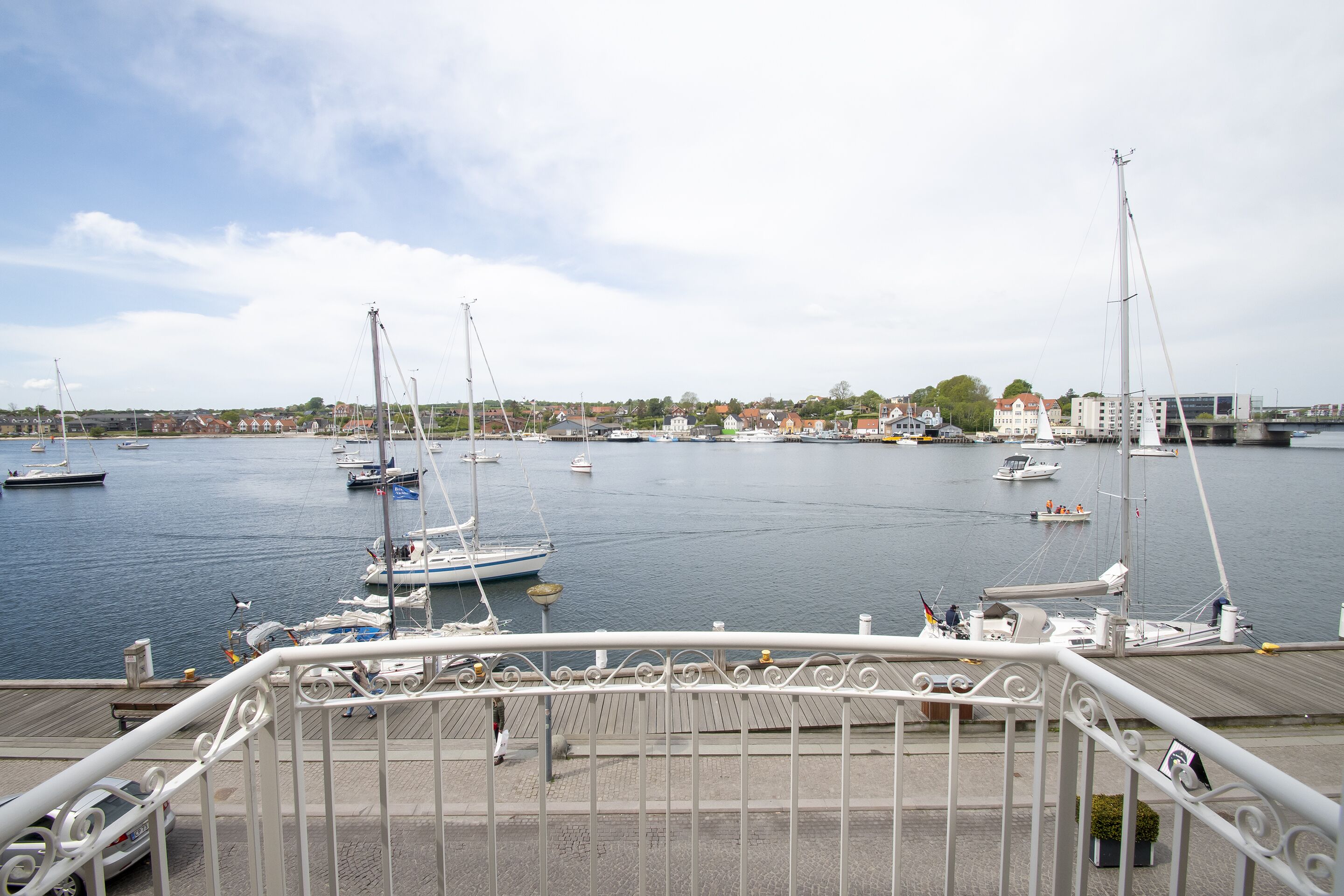 A marina with multiple sailboats docked, a pedestrian walkway, and buildings in the background.