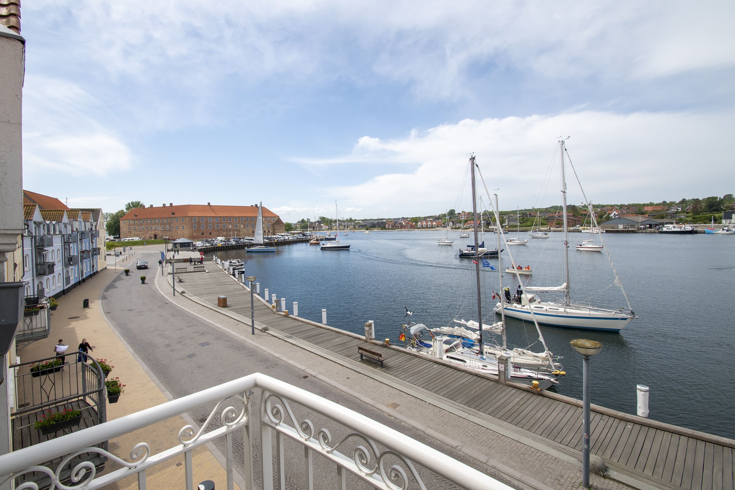 A marina with boats docked, a waterfront promenade, and buildings in the background.