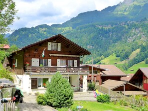 A wooden chalet with a balcony, surrounded by lush greenery and mountains.