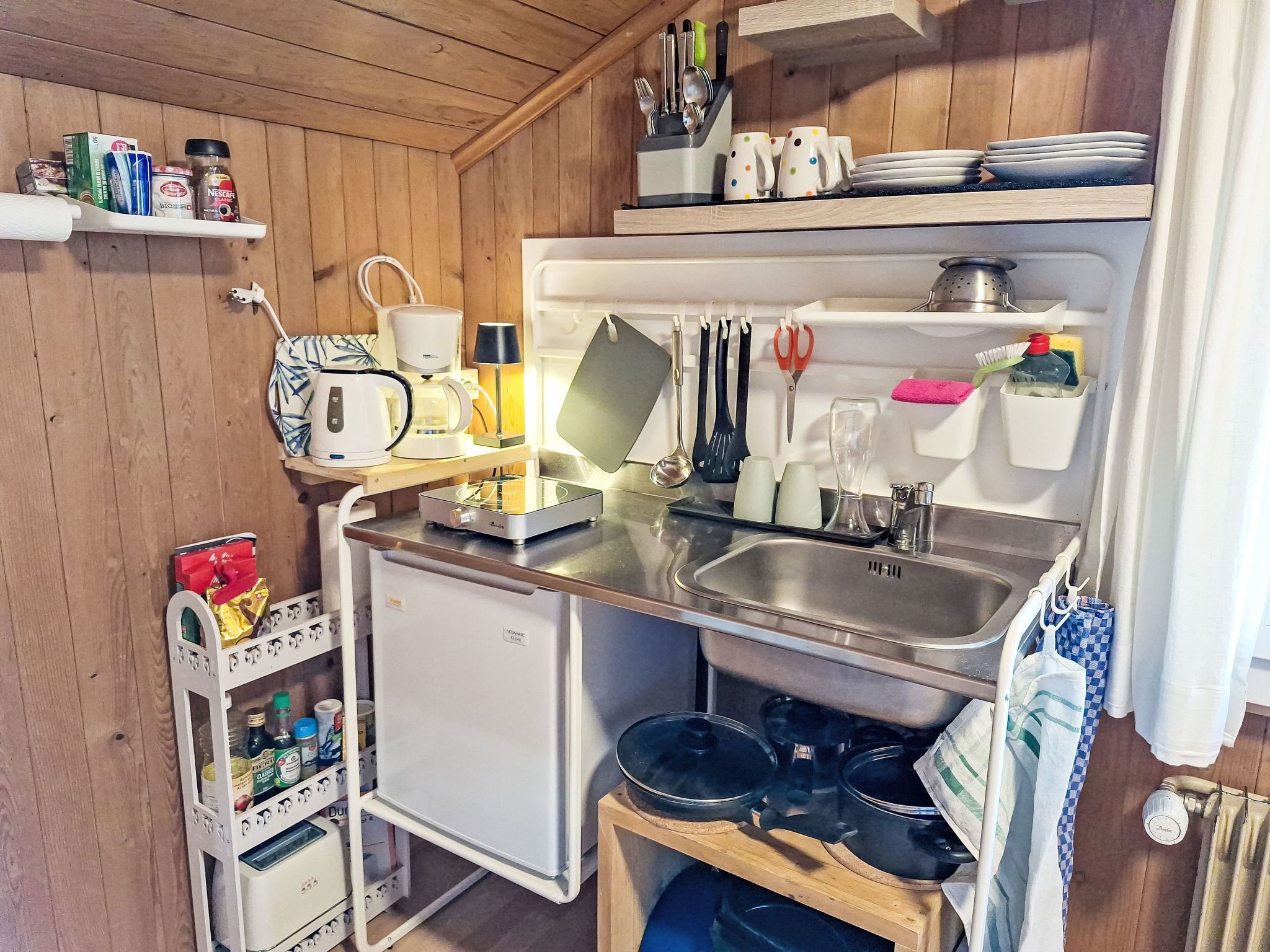 Une cuisine compacte équipée d’un évier, d’un petit réfrigérateur et d’une cuisinière. On y trouve des étagères remplies de divers ustensiles de cuisine et une machine à café.