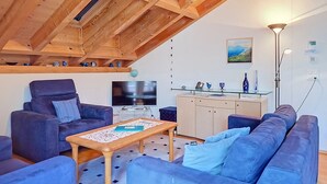 Ein modernes Wohnzimmer mit hoher Decke, Holzbalken, einer Sofagarnitur, einem Couchtisch, einem Fernseher und einer Küchenzeile mit Glasplatte.