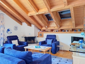 Un salon chaleureux avec un haut plafond en bois, une cheminée et des meubles recouverts d’un tissu bleu.