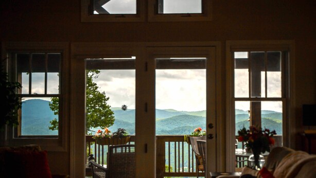 A room with a view of mountains through large windows. There is a chair and a table with a vase of flowers. - Eagle’s Roost - Views from the Top! (Banner Elk)