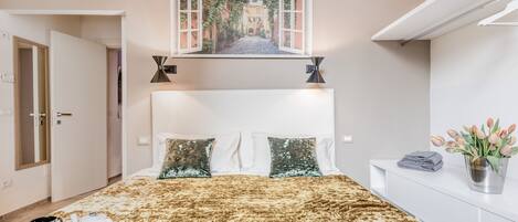 A bedroom with a large bed, a painting of a street view above the bed, a bedside table with a vase of flowers, and a door leading to another room.
