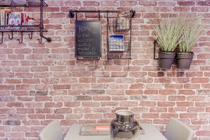 Una pared de ladrillo con un letrero de pizarra, un estante de metal con libros y una postal, y dos plantas en macetas.