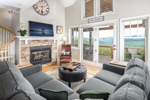 A living room with a fireplace, grey sofas, a round ottoman, a TV, and a view of the outdoors through large sliding doors. - Updated Top Floor Condo! (Banner Elk)