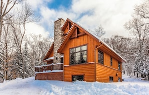 A wooden cabin with a stone chimney, large windows, and a deck.