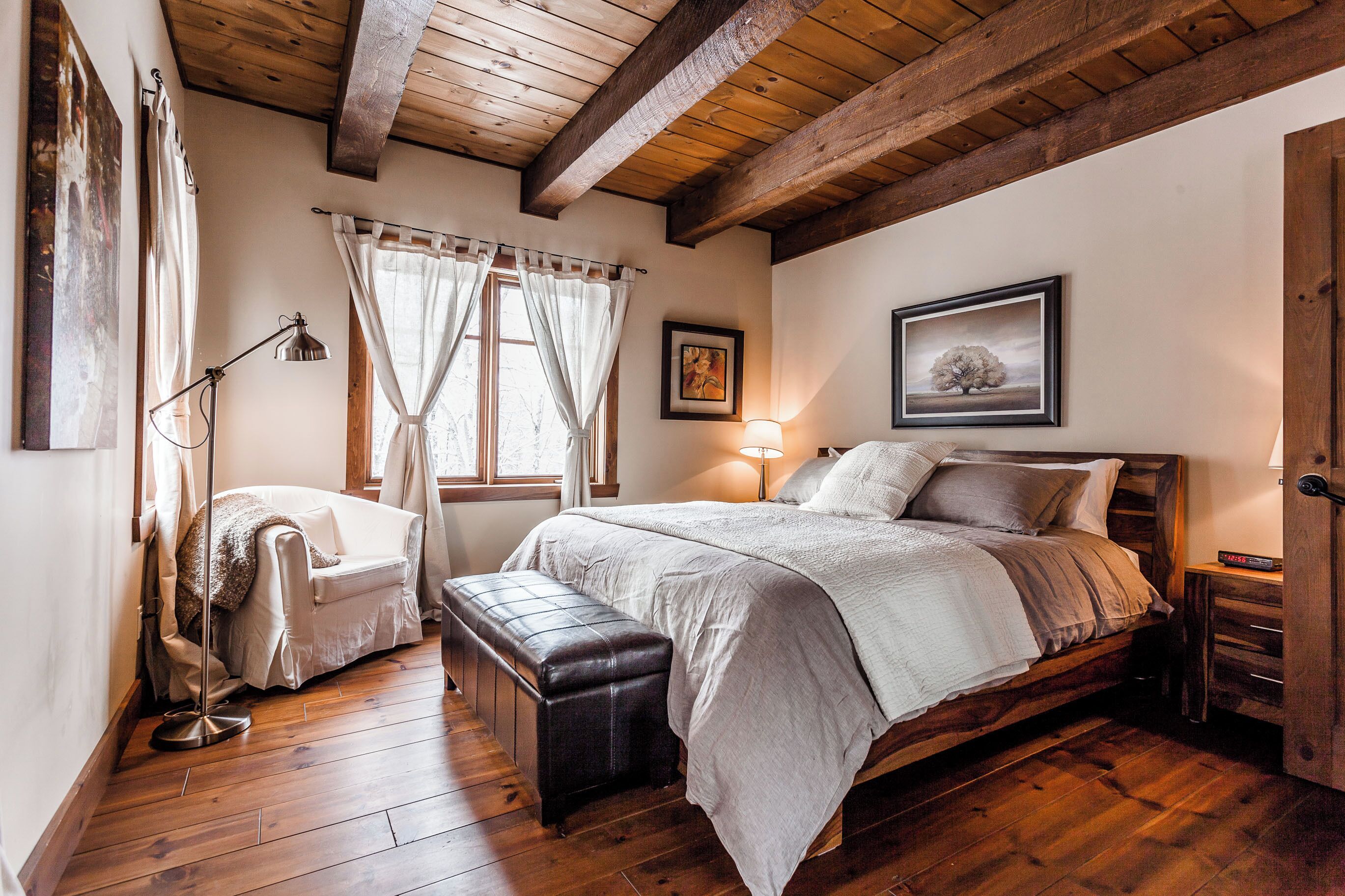 Quarto com cama de madeira, um banquinho, luminária de chão e janela com cortinas.
