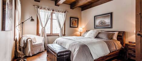Quarto com cama de madeira, um banquinho, luminária de chão e janela com cortinas.