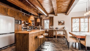 Una cocina con armarios de madera, una isla central, una zona de comedor con mesa y sillas, y un ventanal grande.