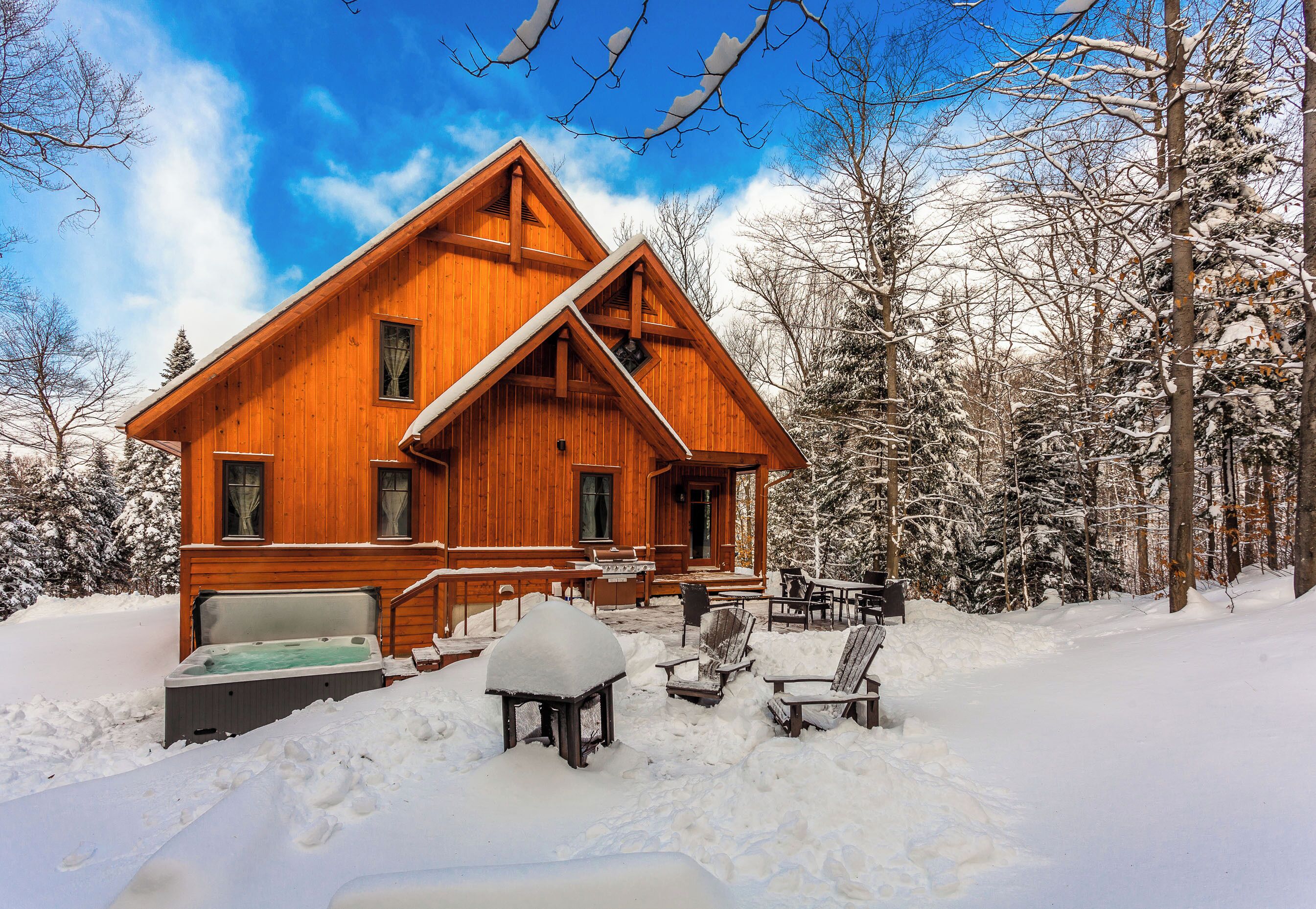 A wooden cabin with a hot tub, outdoor seating, and a snow-covered landscape.