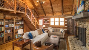 L’intérieur d’un chalet confortable, avec une cheminée, des étagères en bois remplies de livres, un canapé et un escalier.