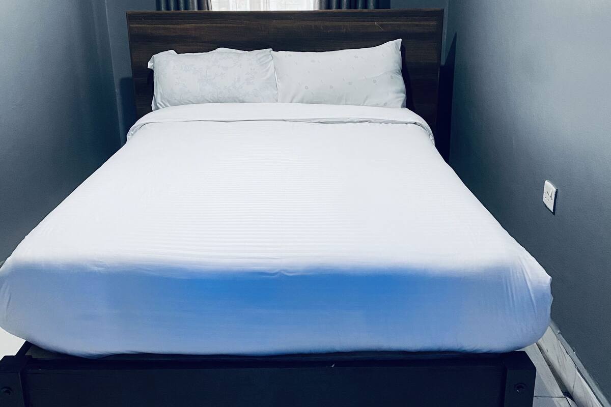A neatly made bed in a room with a wooden headboard, a window with curtains, and a light switch on the wall.