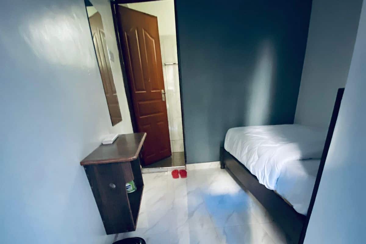 A small, narrow room with a single bed, a wooden cabinet, and a door leading to another room.