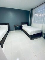 Una habitación de hotel con dos camas individuales, una mesita de noche pequeña y un ventanal con cortinas.