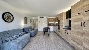 A compact living space with a grey sofa, wooden cabinets, a white door, and a kitchen area with a dishwasher and oven. - Nice studio with balcony located in Fréjus. (Fréjus)