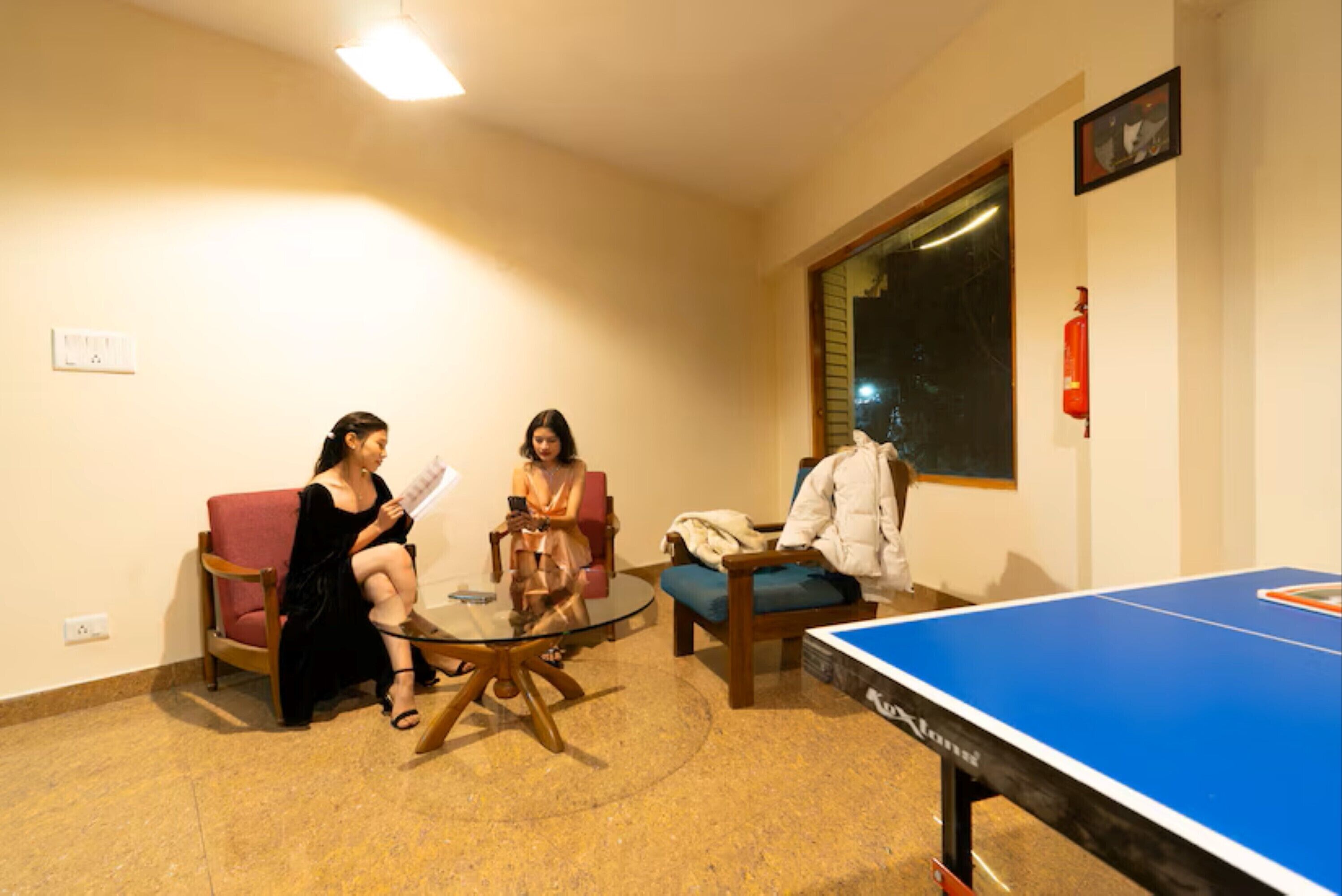 A room with a ping pong table, two chairs, a glass table, and a window.