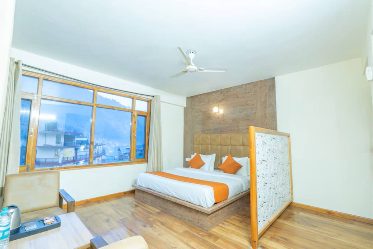 Una habitación de hotel con un ventanal grande, una cama con ropa de cama naranja, suelo de madera y un ventilador de techo.
