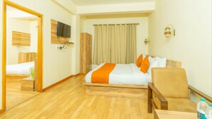 A hotel room with a wooden floor, a bed with white and orange bedding, a chair, a TV mounted on the wall, and a curtain on the window.