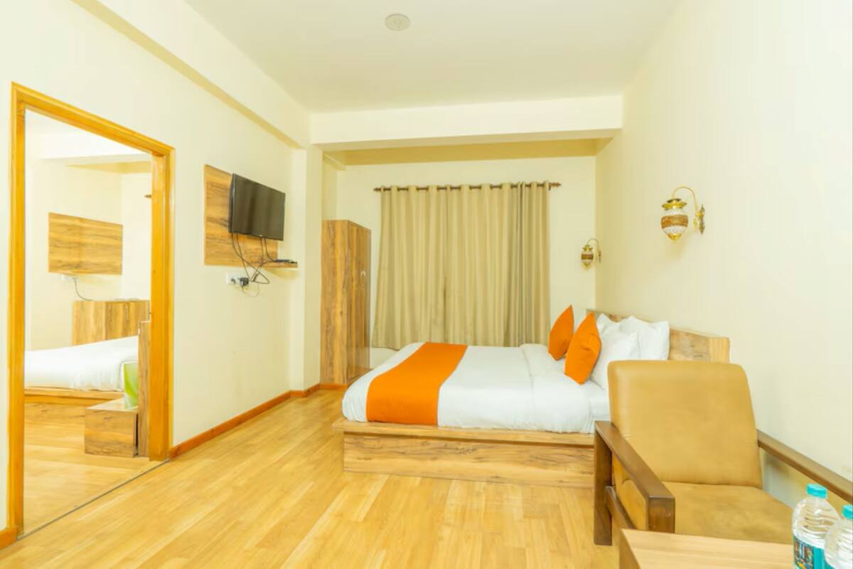 Una habitación de hotel con piso de madera, cama con ropa de cama blanca y naranja, una silla, un televisor montado en la pared y una cortina en la ventana.