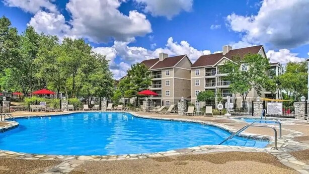 A swimming pool with lounge chairs and umbrellas, surrounded by a building with multiple balconies and trees. - Celebrate Branson (Branson)