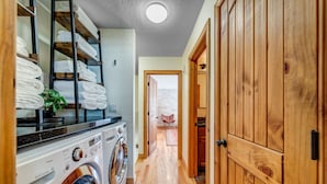 Une buanderie équipée d’un lave-linge et d’un sèche-linge, des étagères en bois où sont pliées des serviettes, et un couloir menant à une pièce avec vue.