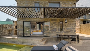 A stone building with a glass door leading to a living area with a sofa and cushions. There is a pool in the foreground and a table with chairs outside. - Lemon House - Valpendre (Valpedre)