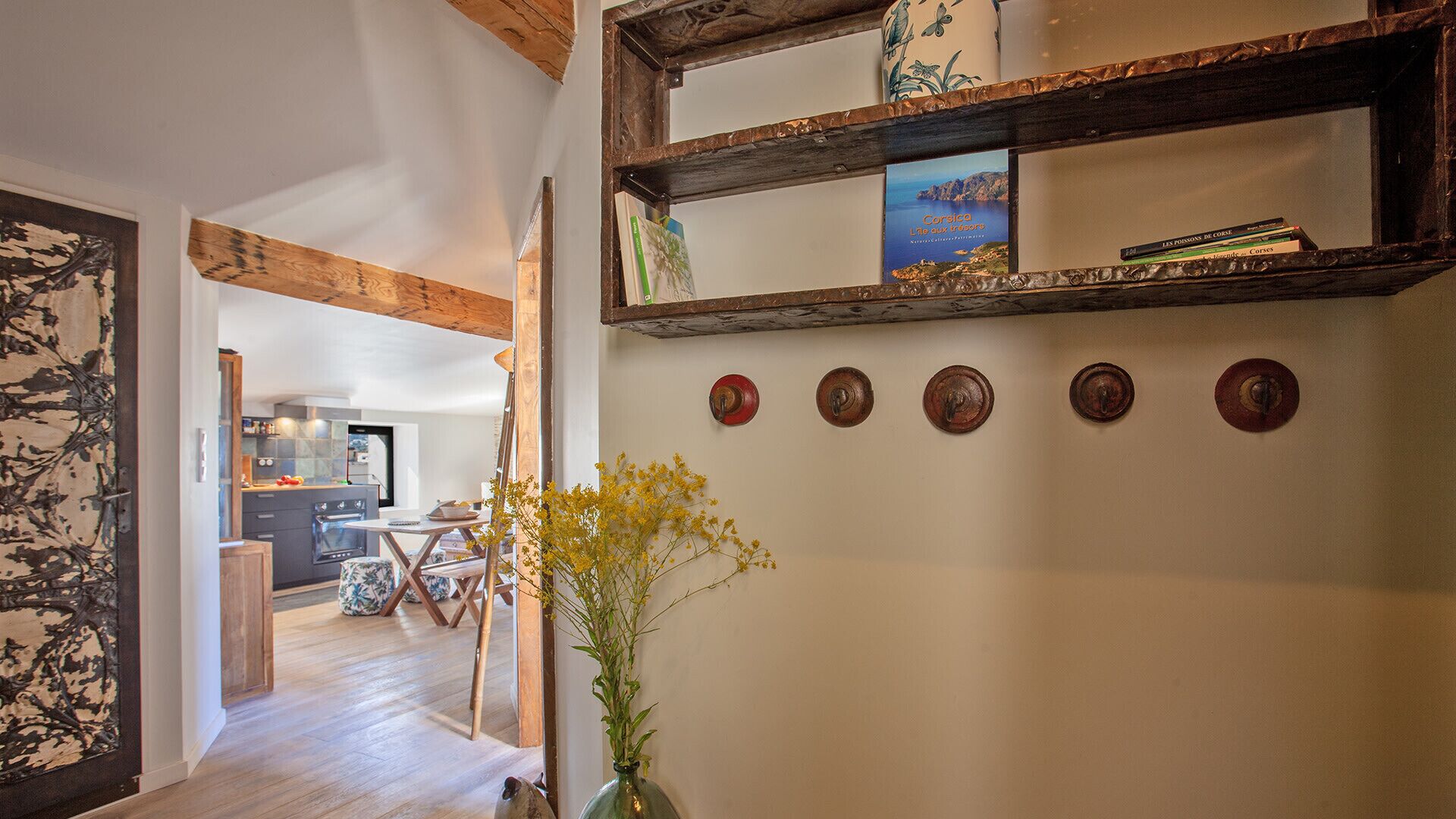 Un couloir avec une étagère en bois rustique où sont exposés des livres et des objets décoratifs, un vase avec des fleurs jaunes et une série d’objets ronds fixés au mur.