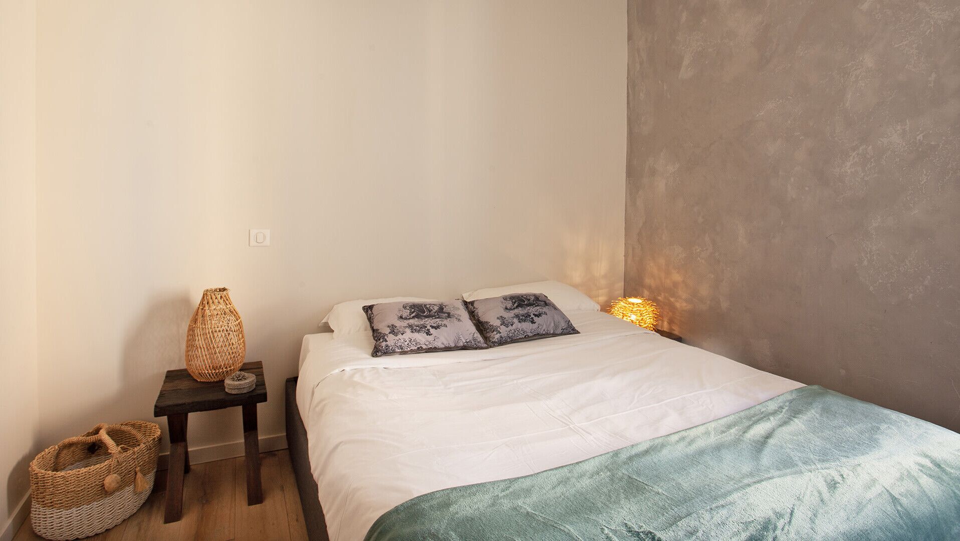 A neatly made bed with white linens and two decorative pillows, a small bedside table with a woven basket, and a wall-mounted lamp.