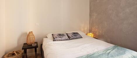 A neatly made bed with white linens and two decorative pillows, a small bedside table with a woven basket, and a wall-mounted lamp.