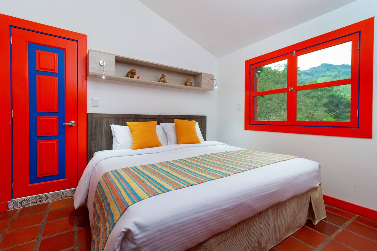 Ein Schlafzimmer mit einem Bett, einem Fenster mit Aussicht, einer roten Tür und einem Regal mit Dekoration.