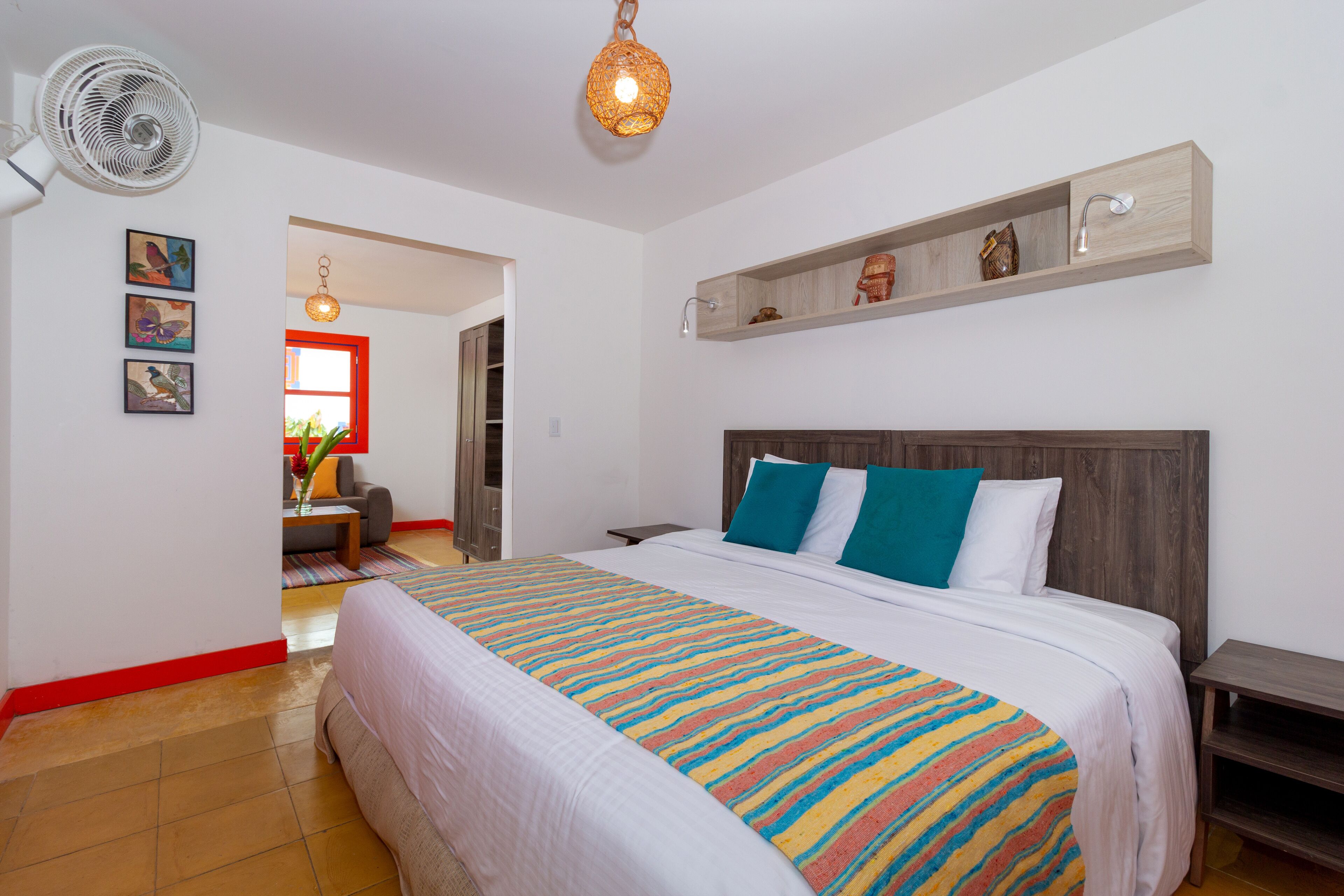 A hotel room with a large bed, a bedside table, a nightstand, a wall-mounted shelf, a ceiling fan, and a door leading to another room.