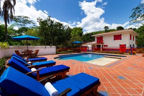 Un espace piscine avec des chaises longues bleues et des parasols, une terrasse carrelée et un bâtiment aux touches de rouge.