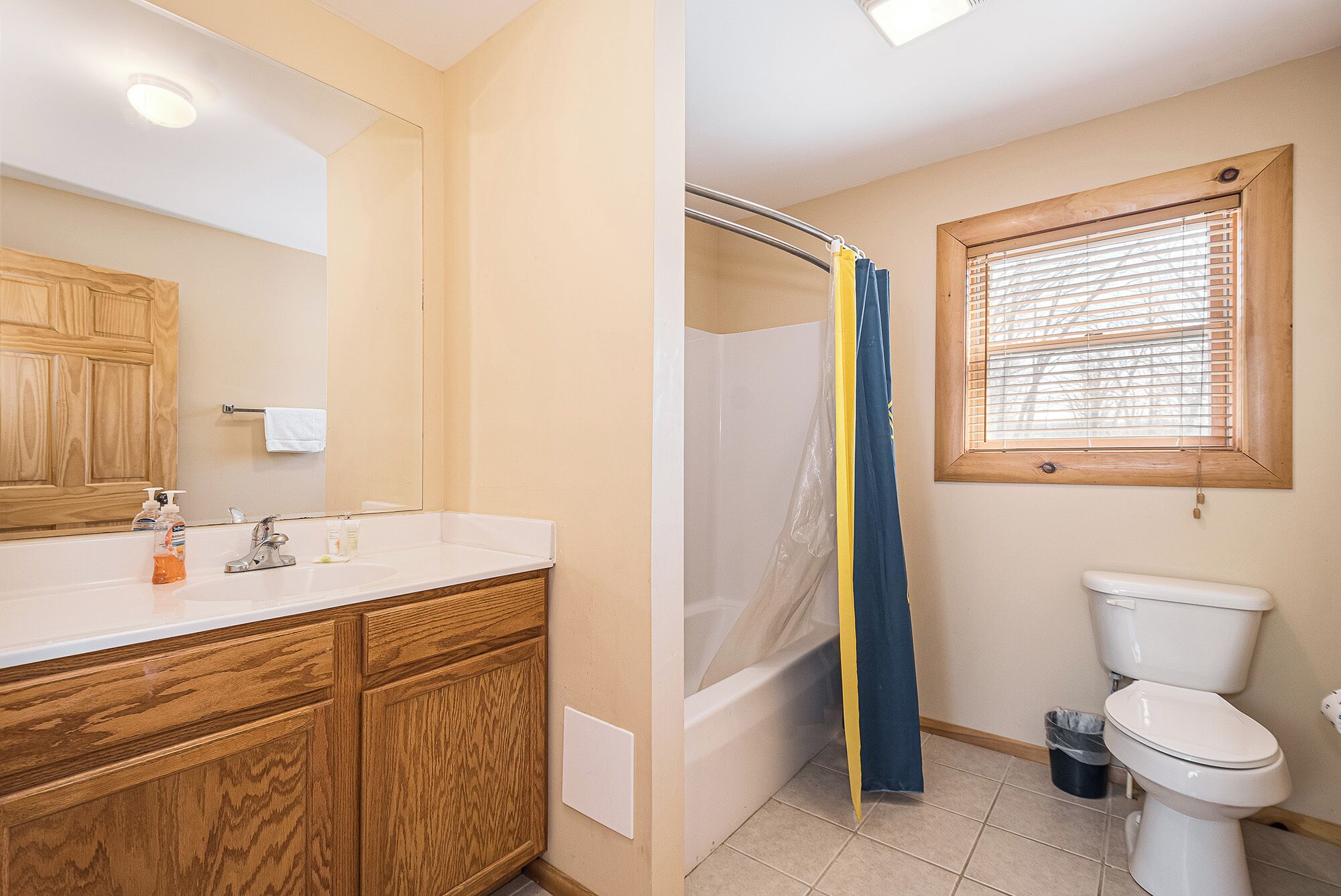 A bathroom with a wooden vanity, a bathtub, a shower curtain, a toilet, and a window with blinds.