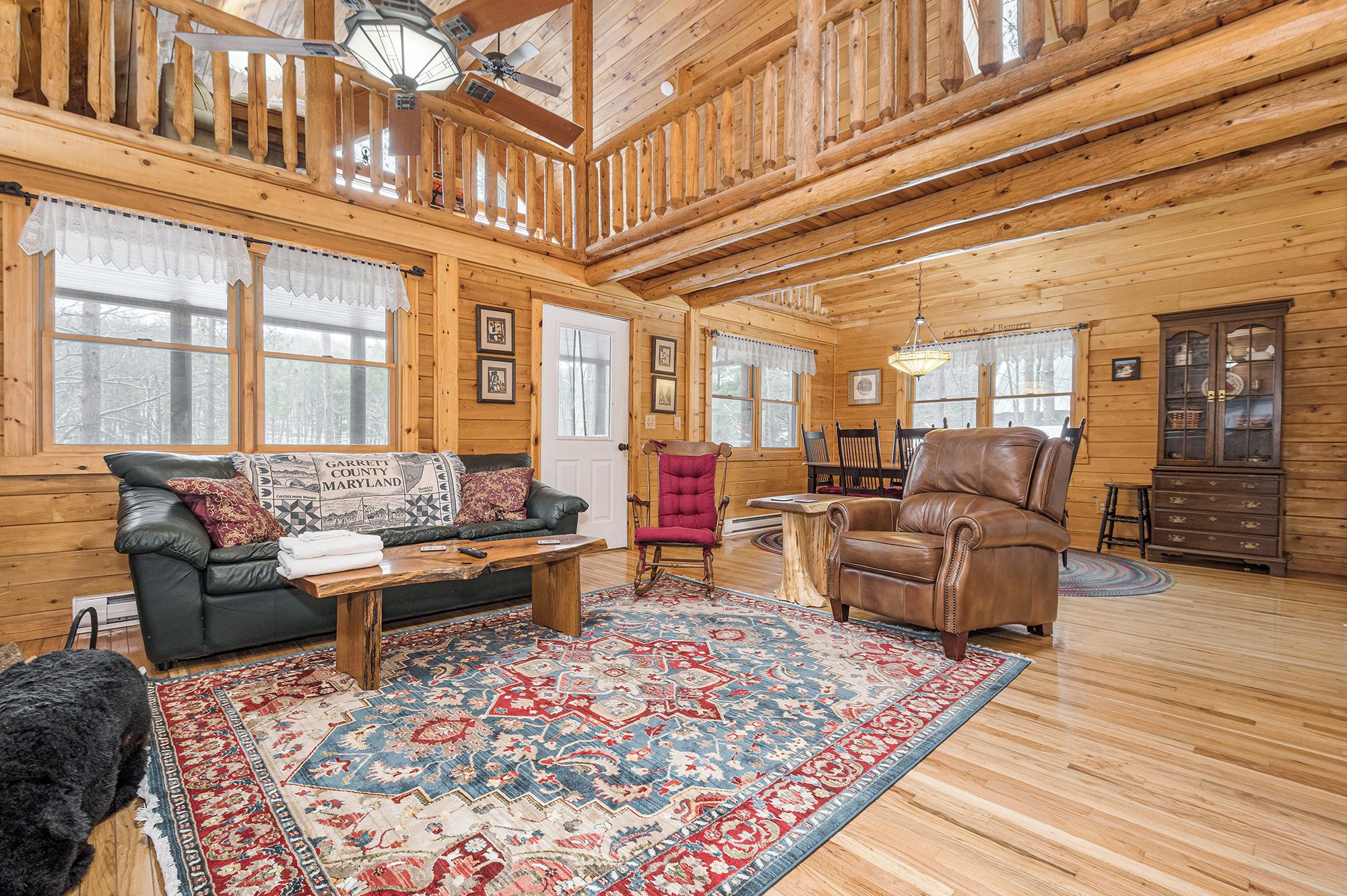 A log cabin interior with a wooden floor, a leather sofa, a red armchair, a wooden coffee table, and a patterned rug.