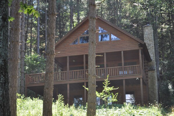 A two-story log cabin with a balcony and a stone chimney, surrounded by tall trees.