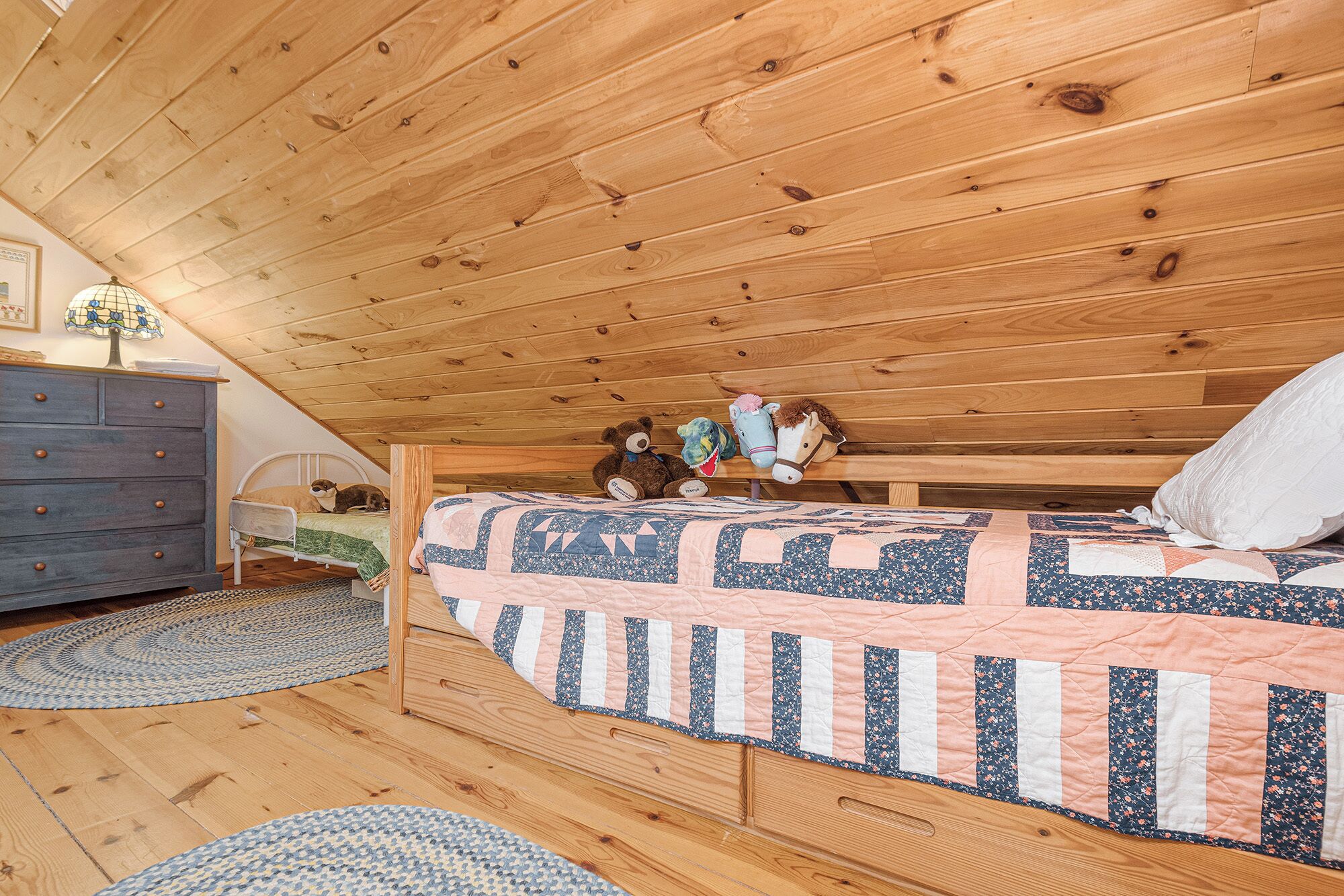 A cozy attic bedroom with a wooden ceiling, a bed with a quilt, a dresser, and a lamp.