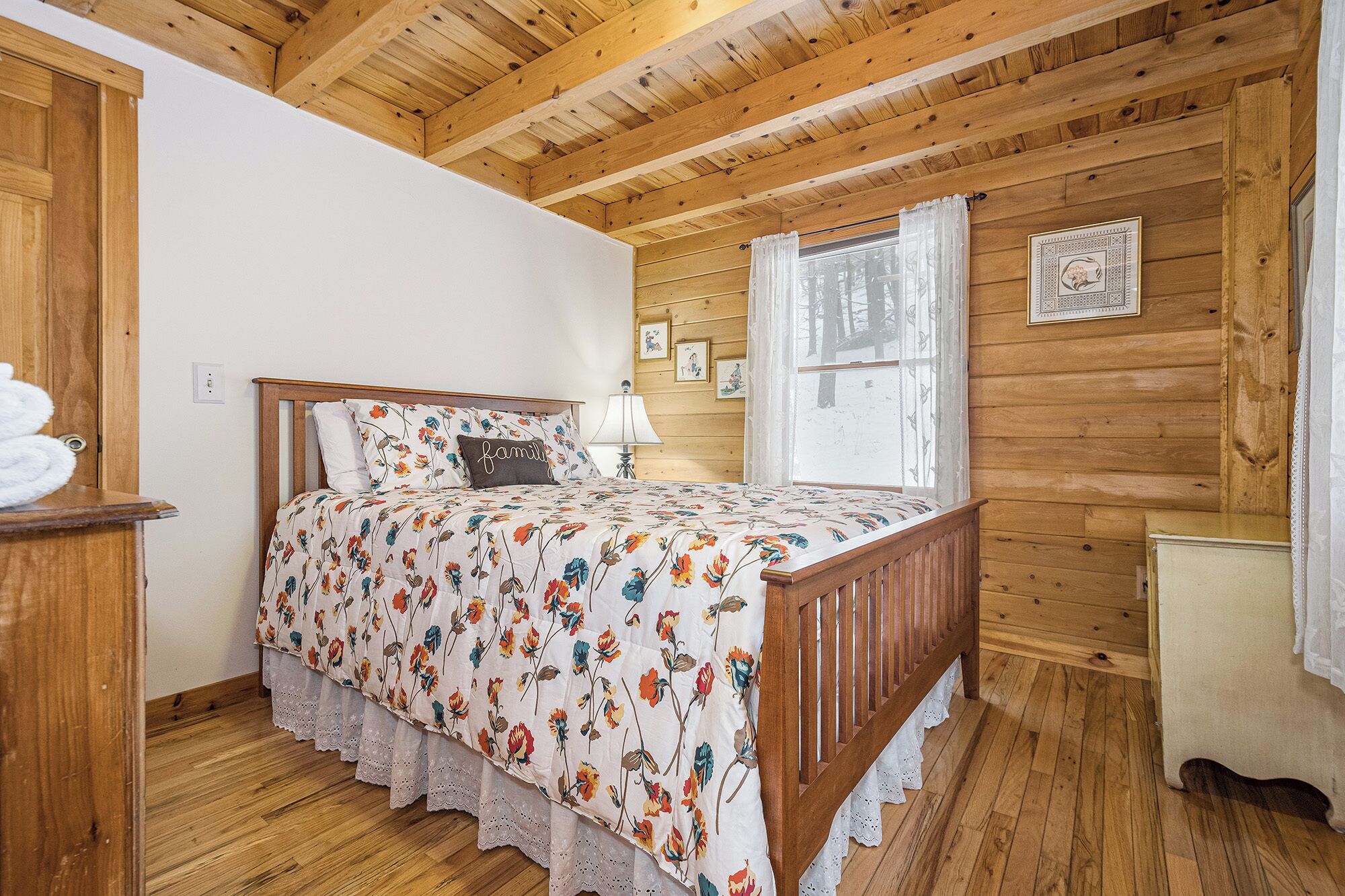 A cozy bedroom with a wooden ceiling, a bed with floral bedding, a bedside table, and a window with curtains.