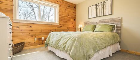 A bedroom with a wooden ceiling, a large window, a bed with green bedding, a nightstand, and a wall-mounted lamp.