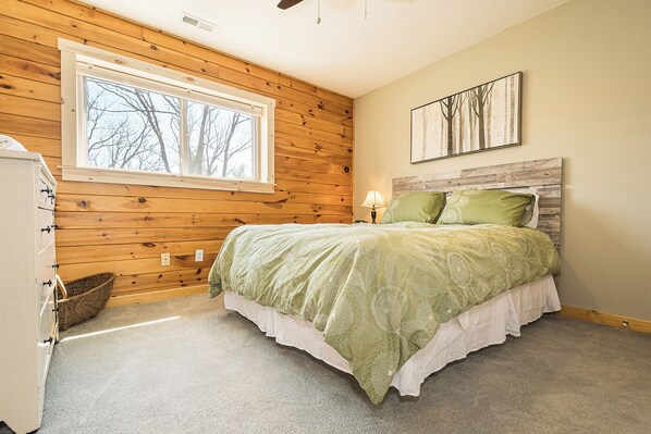 A bedroom with a wooden ceiling, a large window, a bed with green bedding, a nightstand, and a wall-mounted lamp. - Lake Time by Taylor Made Deep Creek (Swanton)