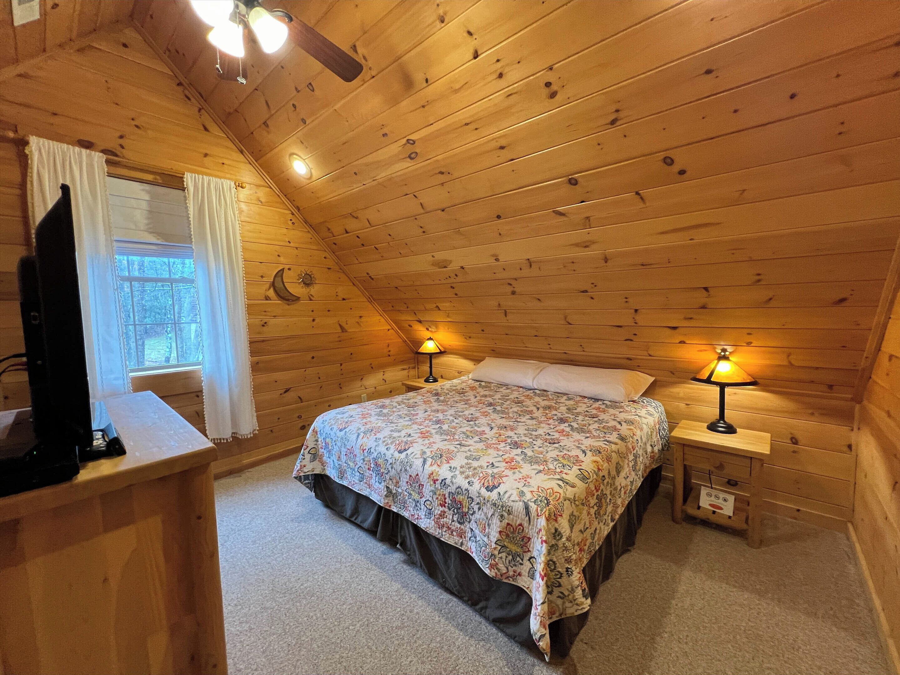 A cozy attic bedroom with a wooden sloped ceiling, a bed with floral bedding, two bedside lamps, and a TV on a wooden dresser.