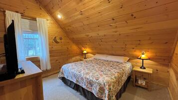 A cozy attic bedroom with a wooden sloped ceiling, a bed with floral bedding, two bedside lamps, and a TV on a wooden dresser.