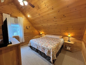 Une chambre confortable située sous les toits, avec un plafond en pente en bois, un lit recouvert d’une literie à motifs floraux, deux lampes de chevet et un téléviseur posé sur une commode en bois.
