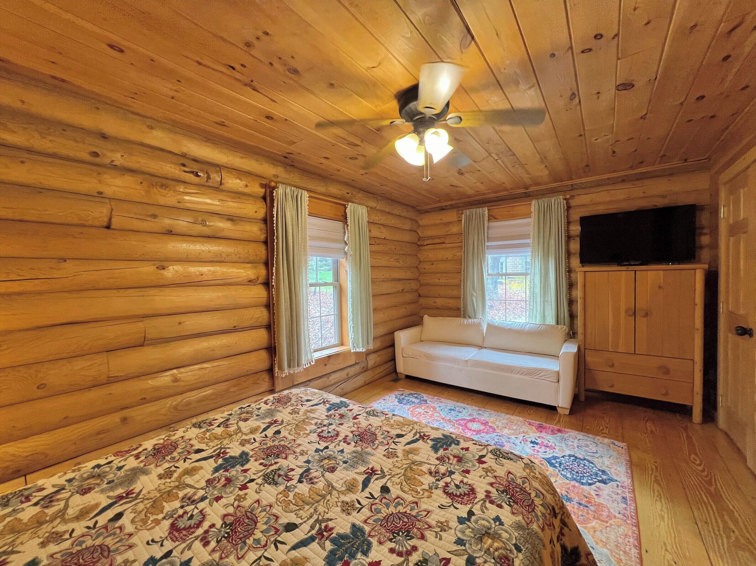Una habitación tipo cabaña de madera con una cama, un sofá, un televisor y un ventilador de techo.