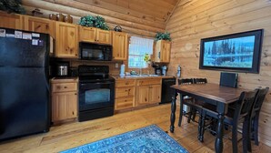 Eine Holzhüttenküche mit Kühlschrank, Herd, Mikrowelle und Holzschränken. Ein Esstisch mit Stühlen und ein Teppichboden sind ebenfalls vorhanden.