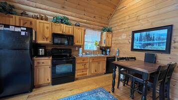 A wooden cabin kitchen with a refrigerator, stove, microwave, and wooden cabinets. There's a dining table with chairs and a rug on the floor.