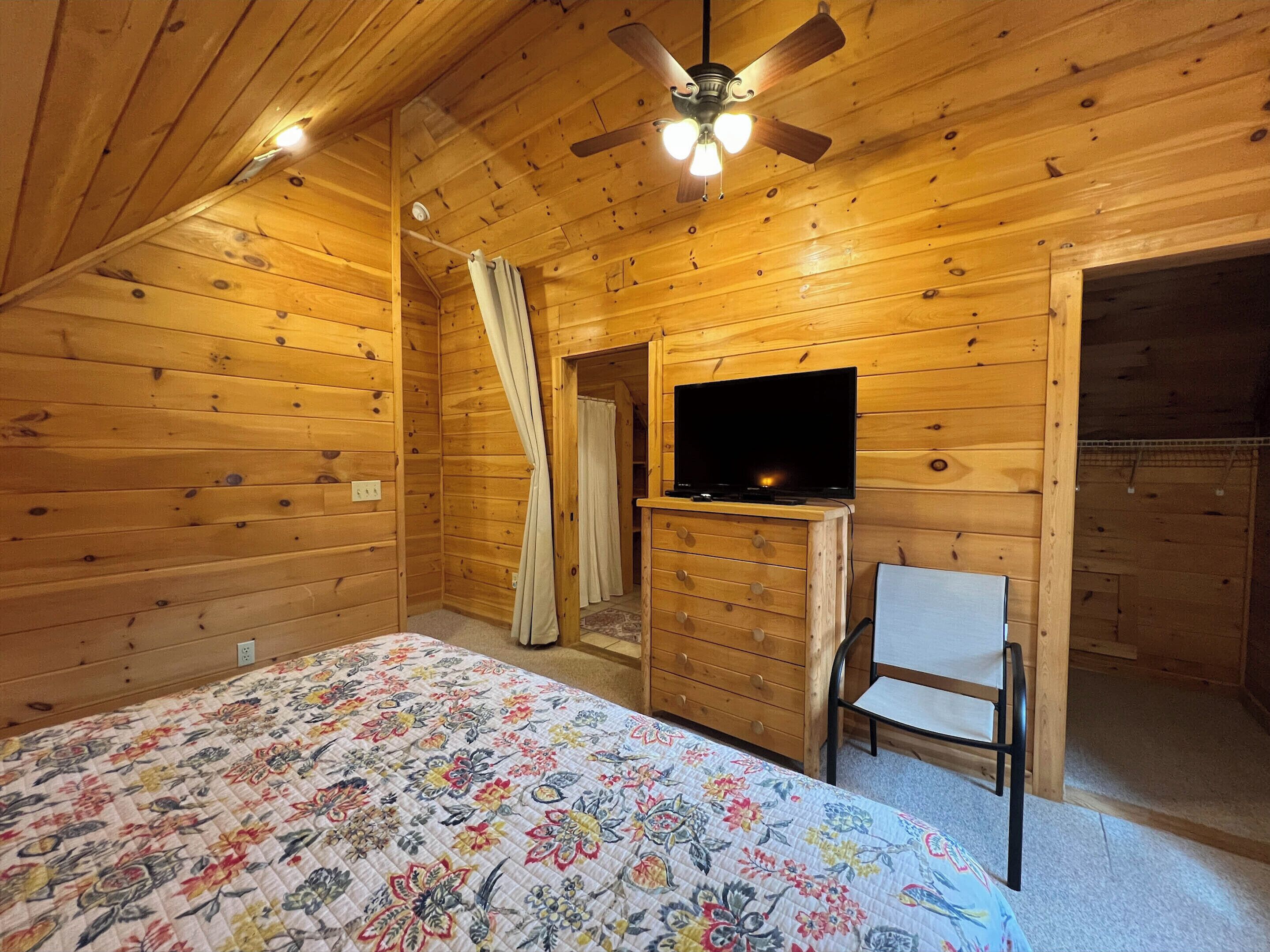 Una habitación de cabaña de madera con una cama, un televisor en una cómoda, un ventilador de techo y una silla.
