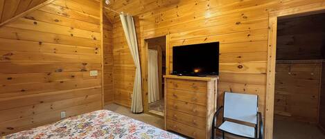Una habitación de cabaña de madera con una cama, un televisor en una cómoda, un ventilador de techo y una silla.