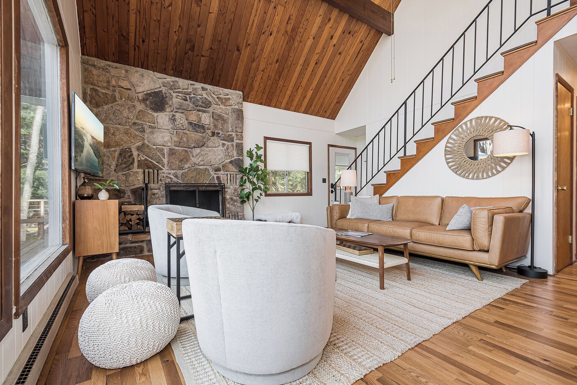 Un amplio salón con chimenea de piedra, una escalera de madera y un ventanal con vistas a una terraza.